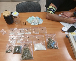 Na stole leżą narkotyki, zapakowane w foliowe torebki strunowe, stoją dwa słoiki, dwie wagi, pieniądze, policjant siedzi obok, sporządza dokumentację