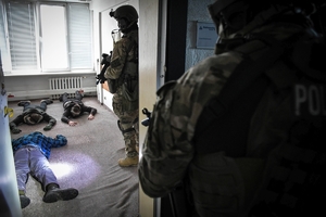 Umundurowani policjanci z bronią, podczas zatrzymywania osób. Na podłodze leżą trzy cywilne osoby, maja ręce założone z karku.