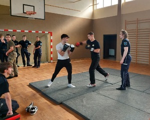 Uczniowie na sali gimnastycznej, podczas pokazu technik interwencji, mają założone rękawie bokserskie, ćwiczą na matach rozłożonych na podłodze sali.