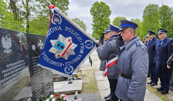 Poczet sztandarowy KPP Zduńska Wola, trzech policjantów stoi przed tablicą, jeden z nich trzyma pochylony sztandar.